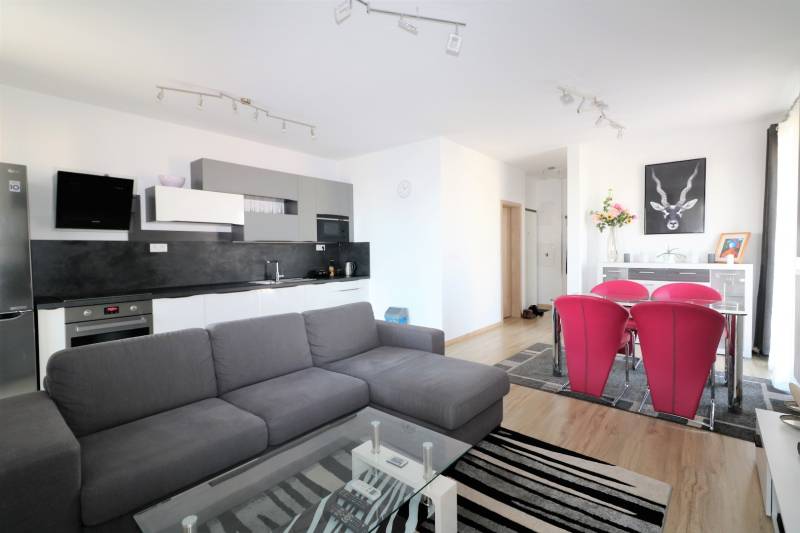 NUERA︱ SLNEČNICE- slnečný 2izbový byt s výhľadom na hrad - Obývacia izba s kuchynským kútom, sivou sedačkou, skleneným stolom a červenými stoličkami.