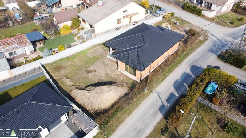 PRIESTRANNÁ 4-IZBOVÁ NOVOSTAVBA 167.38m²- BUNGALOV NA POZEMKU AŽ 702m² - Rodinný dom v Pavliciach pri ceste, so záhradou a susediacimi domami v pozadí.
