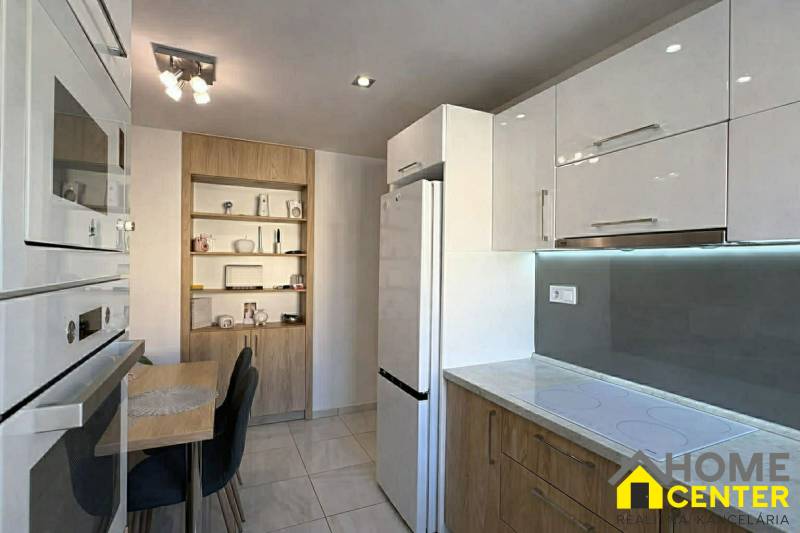 NA PREDAJ vkusne zrekonštruovaný  2 izbový byt, Komárno - Kuchyňa v 2-izbovom byte s bielymi skrinkami, stolom a dreveným dekorom políc.