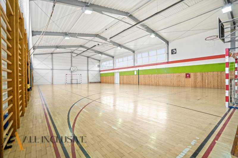 NA PREDAJ ŠKOLSKÁ BUDOVA NA VEĽKOM POZEMKU, MOSTOVÁ - Športová hala s podlahou s dreveným dekorom, basketbalovým košom a sieťkami na oknách.