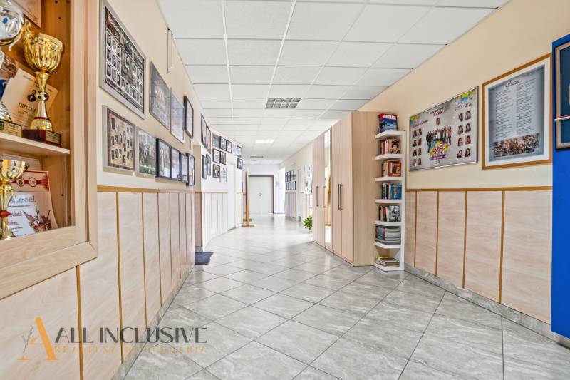 NA PREDAJ ŠKOLSKÁ BUDOVA NA VEĽKOM POZEMKU, MOSTOVÁ - Interiér budovy s trofejami, plagátmi a výstavou fotografií, podlaha s dreveným dekorom.