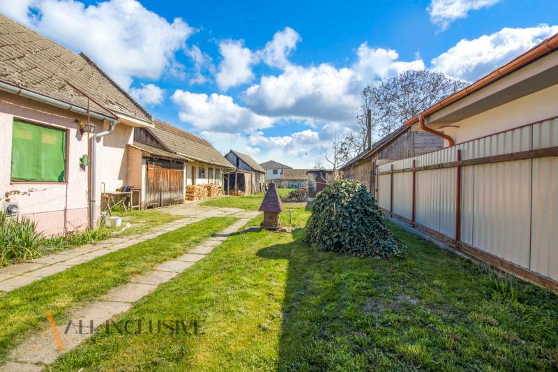 NA PREDAJ 4-IZBOVÝ RODINNÝ DOM NA PRIESTRANNOM POZEMKU - Dvor rodinného domu so záhradou, budovou a stromami na vidieku pod modrou oblohou.