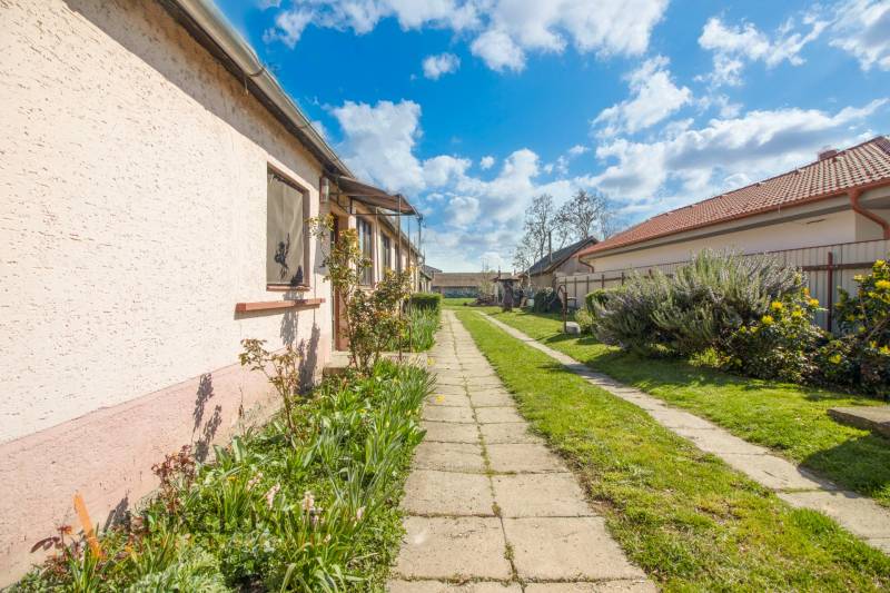 NA PREDAJ 4-IZBOVÝ RODINNÝ DOM NA PRIESTRANNOM POZEMKU - Chodník vedľa rodinného domu s upravenou záhradou a slnečným nebom.