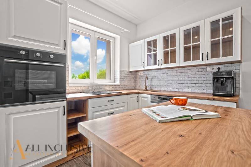 REZERVOVANÉ 3-IZBOVÝ RODINNÝ DOM V OBCI BALOŇ - Kuchynský interiér v Rodinnom dome s podlahou s dreveným dekorom, bielymi skrinkami a oknom.