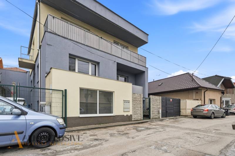 Na predaj 3-izbový byt so záhradkou, Senec - Trojpodlažná budova pri ceste v Senci, obklopená autami a ostatnými budovami. Hotely a penzióny.