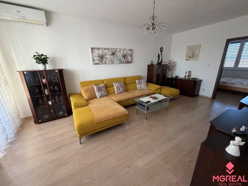 3 izbový byt s balkónom Topoľčany  - Obývacia izba s podlahou s dreveným dekorom, žltou sedačkou a skleneným stolíkom v 3-izbovom byte.
