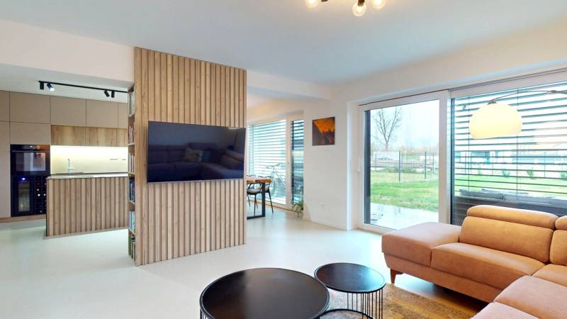 Novozariadený rodinný dom s veľkými spálňami i šatníkom pri Trenčíne   - Obývacia izba s veľkými oknami, pohodlnou sedačkou a dreveným dekorom na stene.