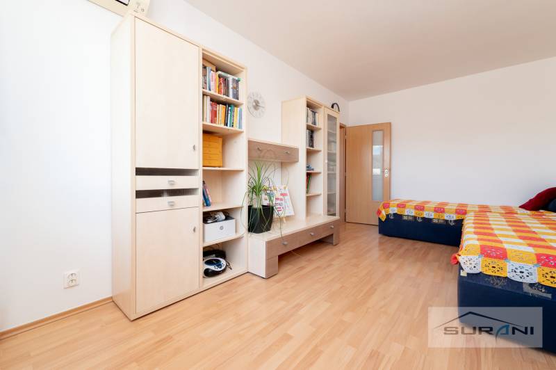 Nové v ponuke | 3-izbový byt 91 m² | 2× balkón | parkovanie | RUSTICA  - Izba s podlahou s dreveným dekorom obsahuje nábytok, regály, dvojlôžko a pestrofarebnú prikrývku.