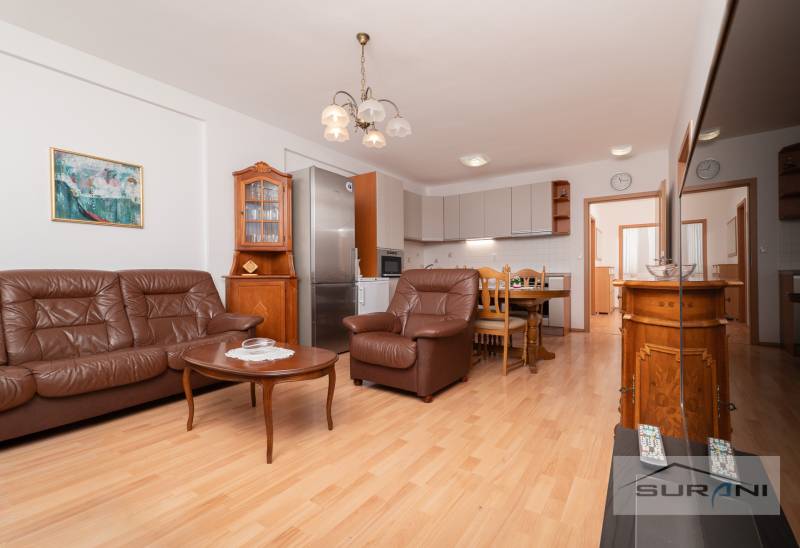 Nové v ponuke | 3-izbový byt 91 m² | 2× balkón | parkovanie | RUSTICA  - Obývačka s koženou sedačkou, jedálenským stolom a kuchyňou, 3-izbový byt, podlaha s dreveným dekorom.