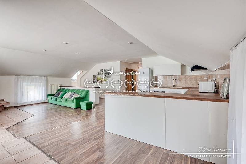 4-izbový podkrovný byt s nebytovým priestorom v obci Pavlice - Obývacia izba s kuchyňou, zelená sedačka, biely nábytok, podlaha s dreveným dekorom.