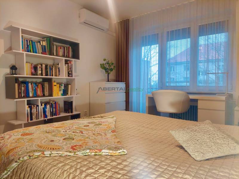 Vkusný, moderný 2 izb, klíma, 870 s ener., Sibírska ul./Nové Mesto - Spálňa v 2-izbovom byte s posteľou, poličkami plnými kníh a pracovným stolom.