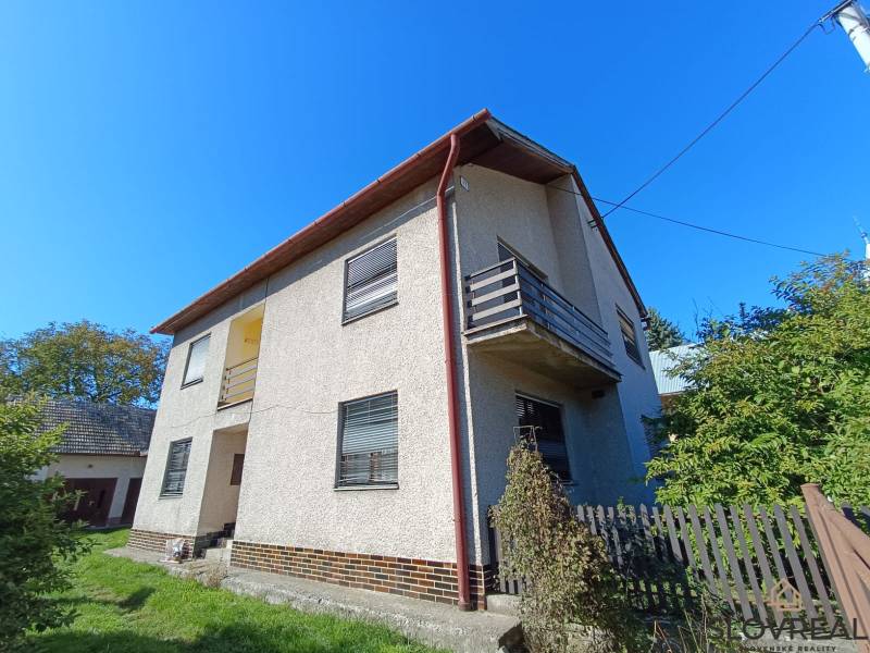 : Predaj rodinného domu s veľkým pozemkom v obci Pitelová  - Rodinný dom v Pitelovej s balkónom a záhradou, modrá obloha, plot a zeleň.