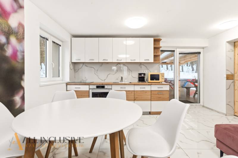 NADŠTANDARDNÁ REKREAČNÁ CHATA | DUCHONKA  - Kuchyňa v chate s bielym stolom, stoličkami a dreveným dekorom na skrinkách.