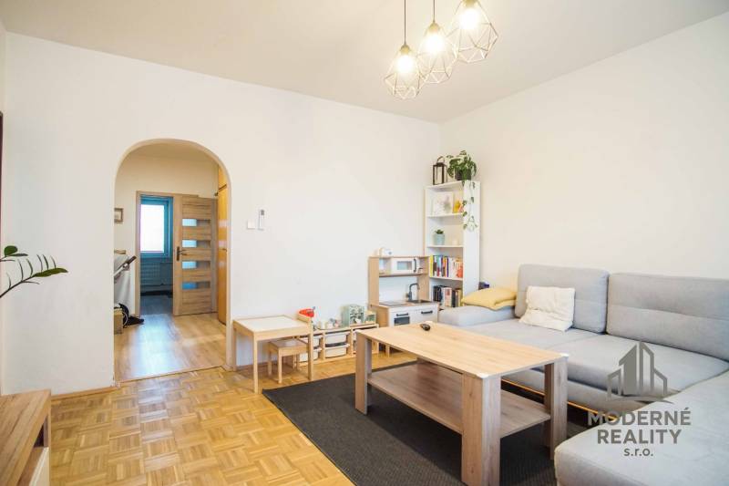 3-izbový byt s loggiou na sídlisku JUH, Nové Zámky – 61,37m2 - Obývacia izba v 3-izbovom byte s podlahou s dreveným dekorom a svetlým nábytkom.