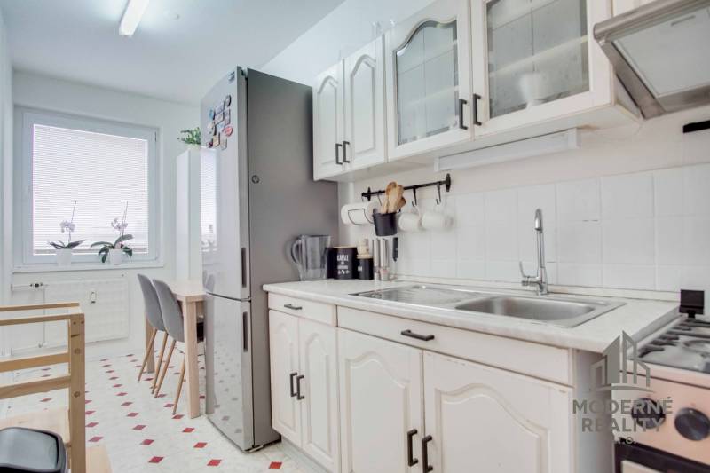 3-izbový byt s loggiou na sídlisku JUH, Nové Zámky – 61,37m2 - Kuchyňa v 3-izbovom byte s bielymi skrinkami, jedálenským stolom a veľkou chladničkou.