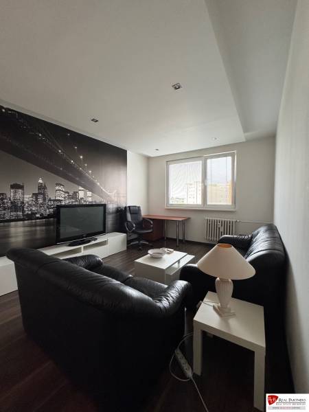 Prenájom 2-izb bytu v Bratislave-Rača - Obývacia izba s čiernou sedačkou, TV, obrazom mesta a pracovným stolom v 2-izbovom byte.