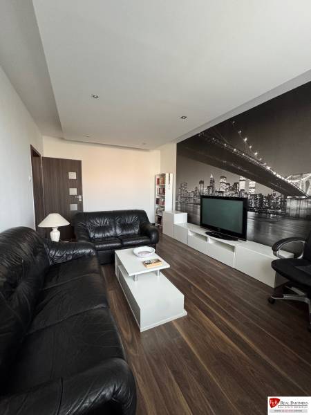 Prenájom 2-izb bytu v Bratislave-Rača - Obývačka 2-izbového bytu s čiernymi pohovkami, podlahou s dreveným dekorom a veľkou tapetou.