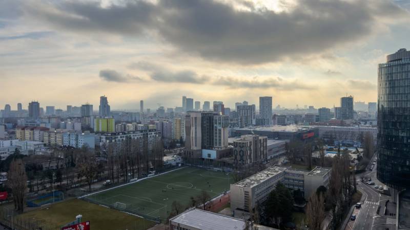 I na predaj 2i byt v komplexe Tri Veže na Bajkalskej - Panoráma Bratislavy z Bajkalskej, ukazuje mestskú krajinu s budovami a športovým ihriskom.