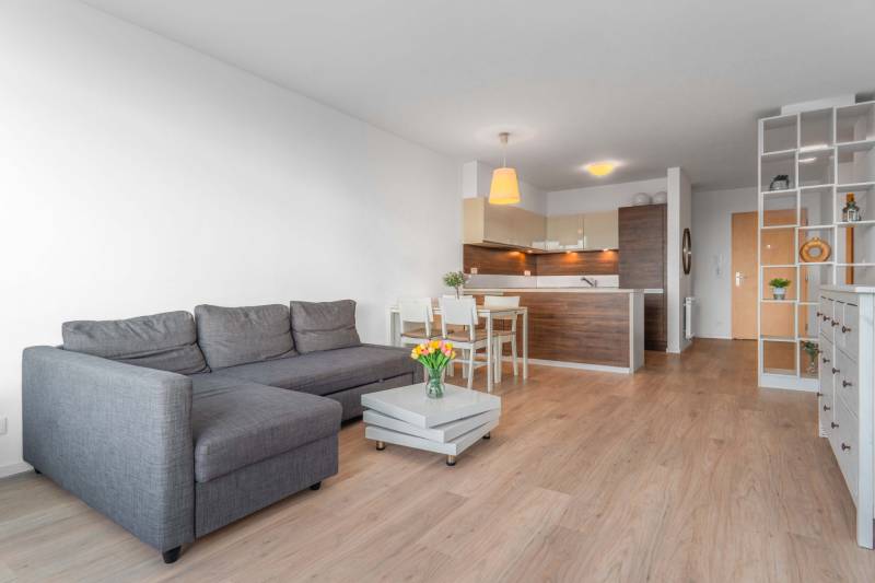 I na predaj 2i byt v komplexe Tri Veže na Bajkalskej - Obývacia izba v 2-izbovom byte s podlahou s dreveným dekorom, sivou sedačkou a kuchyňou.