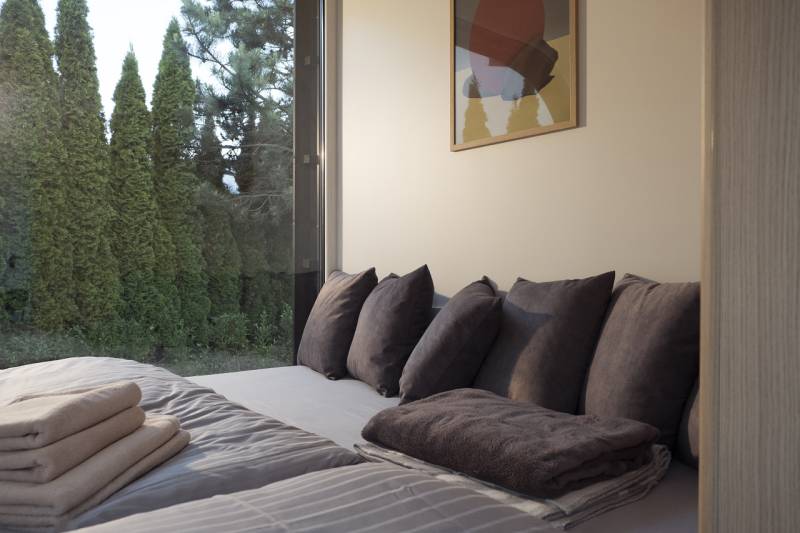 I Predaj Ecocapsule – autonómne ekologické bývanie - Pohodlná posteľ so sivými vankúšmi a výhľadom na prírodu cez veľké okno.