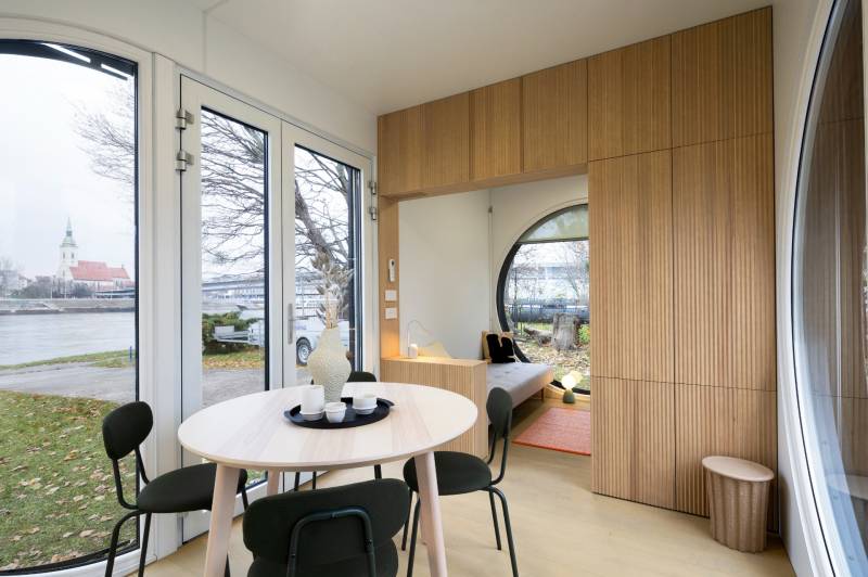 I Predaj Ecocapsule – autonómne ekologické bývanie - Interiér so stolom, stoličkami, podlahou s dreveným dekorom, výhľadom na rieku a kostol.