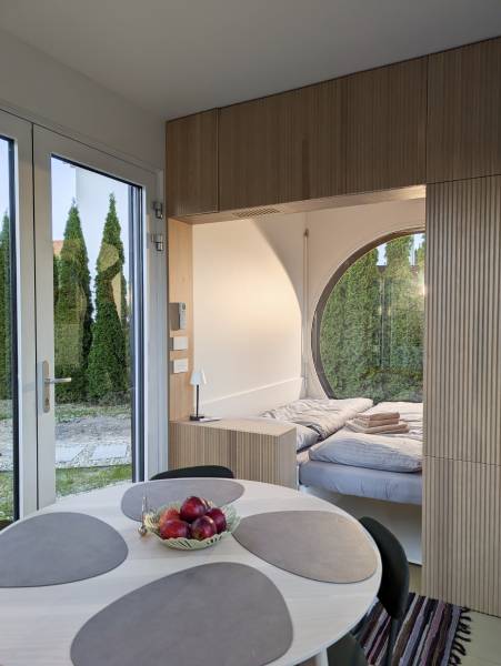 I Predaj Ecocapsule – autonómne ekologické bývanie - Interiér so stolom, ovocím a posteľou pri obloku, podlaha s dreveným dekorom.