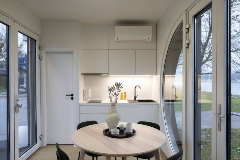 I Predaj Ecocapsule – autonómne ekologické bývanie - Interiér s bielou kuchynkou, okrúhlym stolom a veľkými oknami, podlaha s dreveným dekorom.