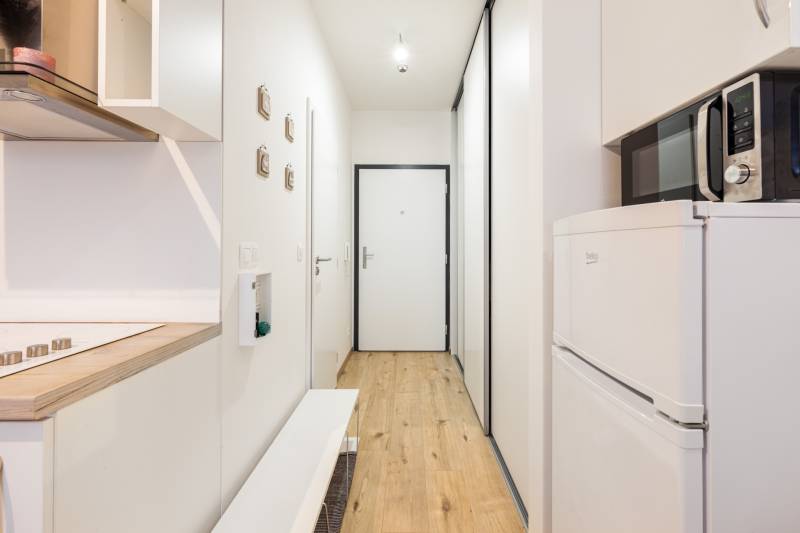 Prenájom 1-izbového bytu, Béžová, Béžová, Petržalka - REZERVOVANÉ - Úzka chodba s kuchynskou linkou, mikrovlnkou a podlahou s dreveným dekorom v 1-izbovom byte.