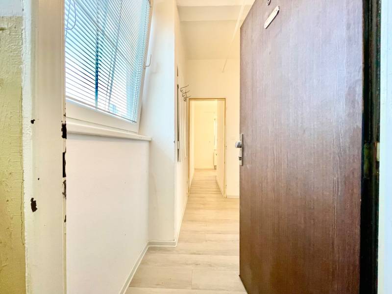 PREDAJ 1 - izbový byt, Bratislava - Ružinov, ul. Trenčianska - Chodba s podlahou s dreveným dekorom, biele steny, veľké okno s žalúziami.