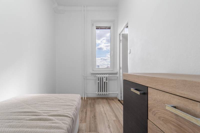 Komfortný 1,5-izbový byt s balkónom v Ružinove na predaj - Jednoduchá izba s posteľou, komodou a podlahou s dreveným dekorom v 1-izbovom byte.