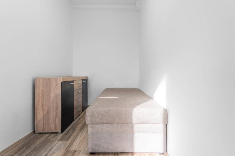 Komfortný 1,5-izbový byt s balkónom v Ružinove na predaj - Úzka miestnosť s posteľou a komodou, podlaha s dreveným dekorom, 1-izbový byt.