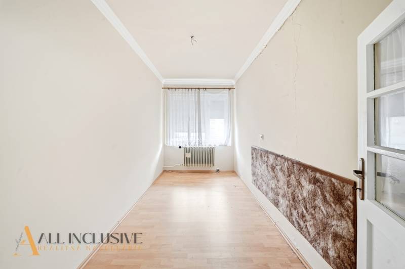 Na predaj 4-izbový rodinný dom, Topoľníky - Úzka izba s podlahou s dreveným dekorom, okno so záclonami, radiátor.