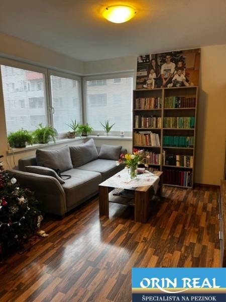 ÚTULNÝ 2 IZBOVÝ BYT - SILVANIUM  - Obývačka v 2-izbovom byte so sedačkou, knihovnicou a stolom. Veľké okná dodávajú svetlo.