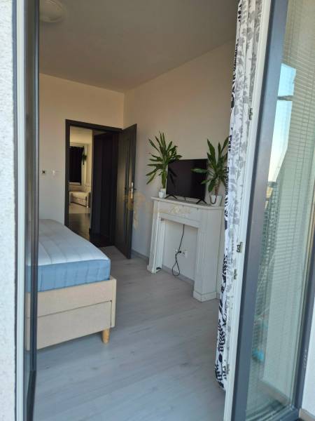 3-izb. byt v obci Blatná na Ostrove, balkón, lodžia, novostavba - Spálňa s posteľou, televízorom a rastlinami v 3-izbovom byte s podlahou s dreveným dekorom.