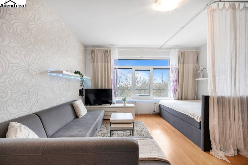 Na predaj príjemný zariadený 1 izbový byt na ulici Antolská, Petržalka - Obývacia izba s pohovkou, závesom a televíziou, podlaha s dreveným dekorom.