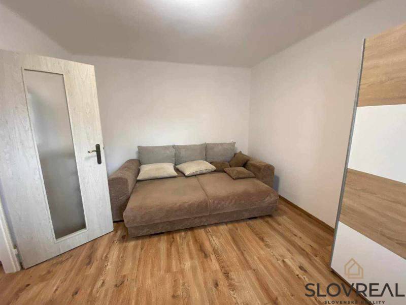 Predaj 3 - izbového bytu - Nová Baňa - Izba s hnedou pohovkou, bielymi stenami a podlahou s dreveným dekorom v 3-izbovom byte.
