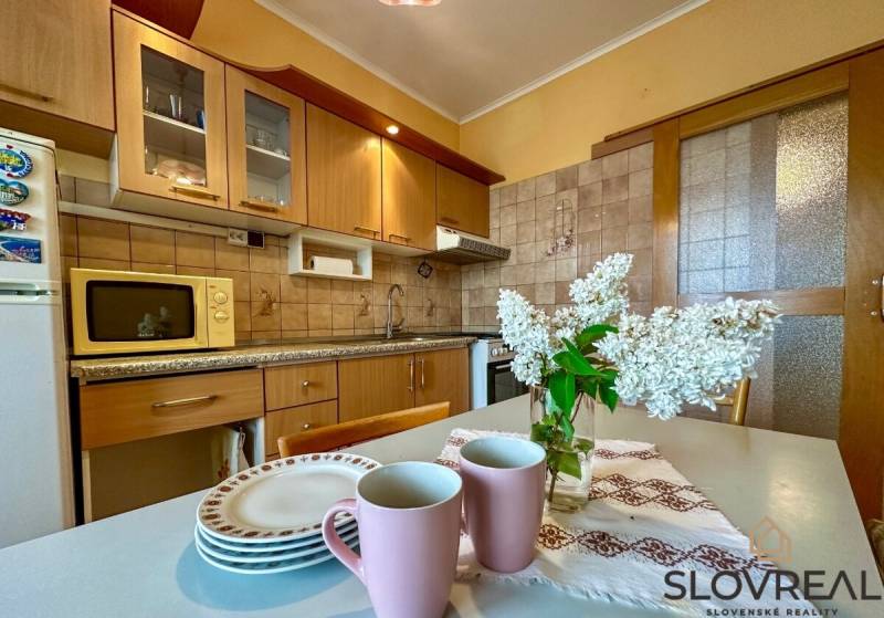 Predaj 3 - izbového bytu - Nová Baňa - Kuchyňa v 3-izbovom byte s drevenými skrinkami, kvetmi a riadom na stole.