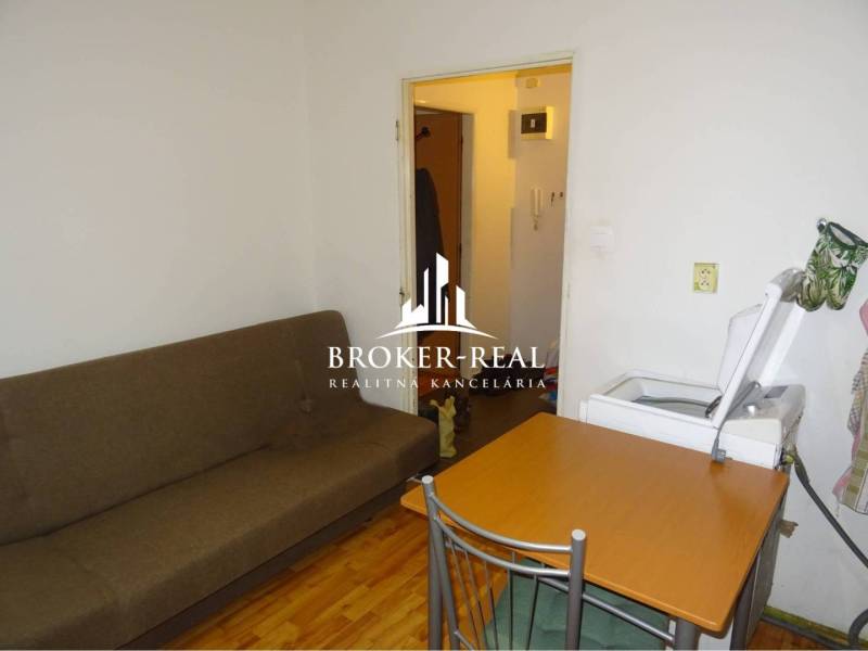 1 - izbový byt Košice, Popradská - Obývacia izba v 1-izbovom byte s gaučom, stolom a práčkou, podlaha s dreveným dekorom.