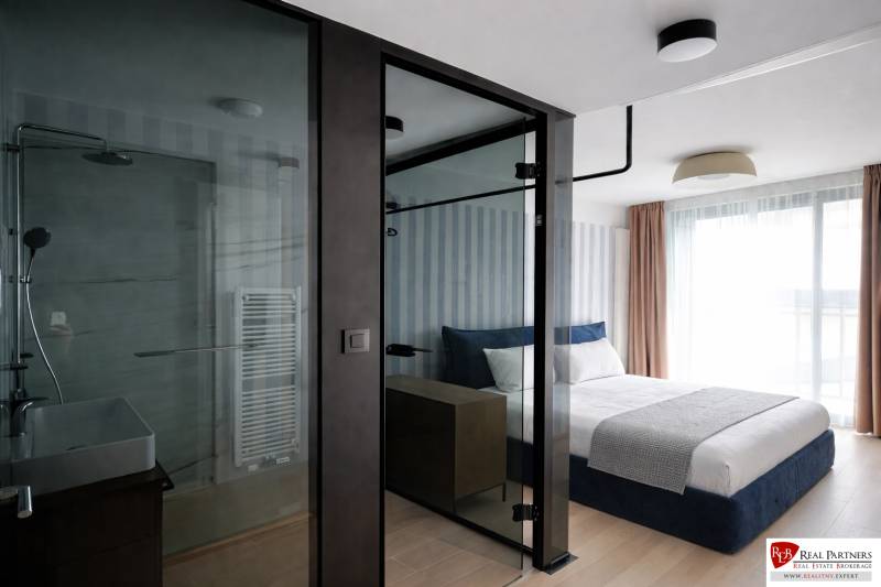 Prenájom 2-izbový byt s loggiou,55m2, EUROVEA TOWER - Spálňa s presklenou kúpeľňou, posteľou a podlahou s dreveným dekorom v 2-izbovom byte.