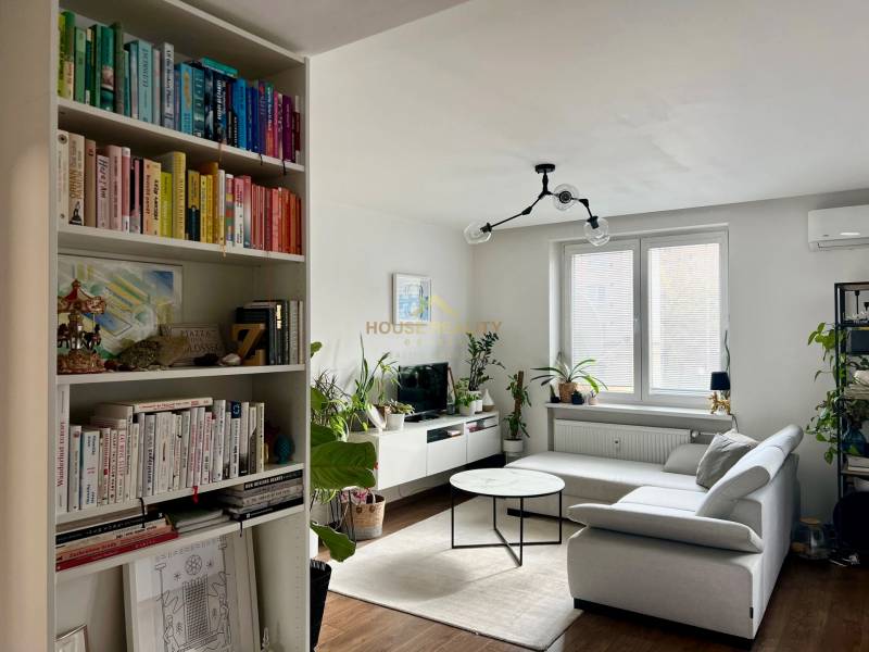 Prenájom 2,5 izbový byt, Trenčianska ulica, Bratislava II. Ružinov - Obývačka v 2-izbovom byte s knihovnicou, pohovkou a oknom, podlaha s dreveným dekorom.