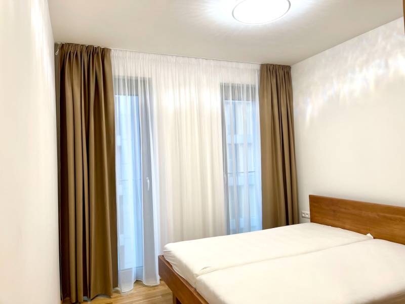 I 3 i byt s parkovaním v rezidenčnom komplexe METROPOLIS - Spálňa s posteľou v 3-izbovom byte, posteľ s dreveným dekorom a závesmi.