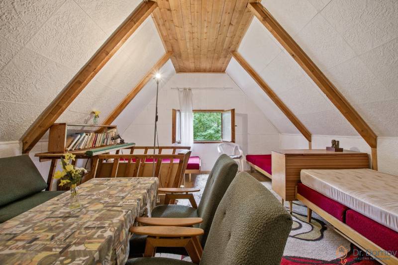 NA PREDAJ CHATA V REKREAČNEJ OBLASTI RUDAVA, OBEC MALÉ LEVÁRE - Podkrovie chaty s dreveným dekorom, stolom, pohovkou a posteľami pre útulnú atmosféru.