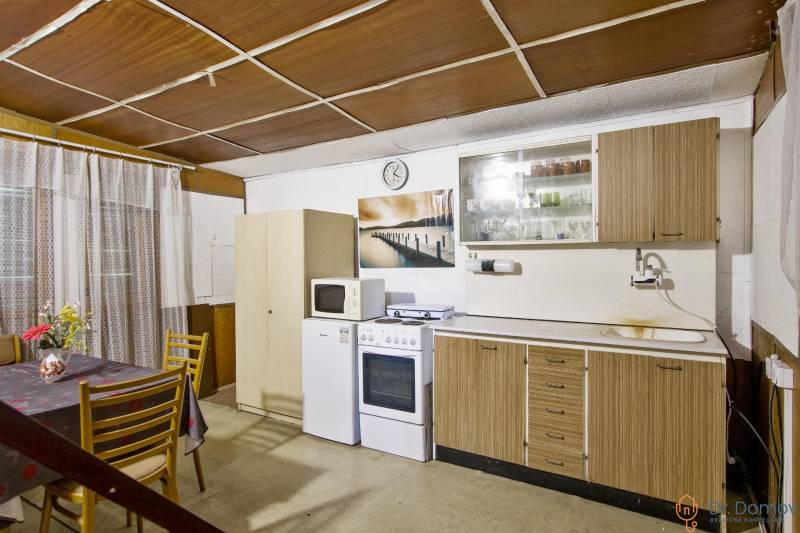 NA PREDAJ CHATA V REKREAČNEJ OBLASTI RUDAVA, OBEC MALÉ LEVÁRE - Kuchyňa chaty s oknom a kuchynskou linkou, pričom stôl má kvetinovú výzdobu.