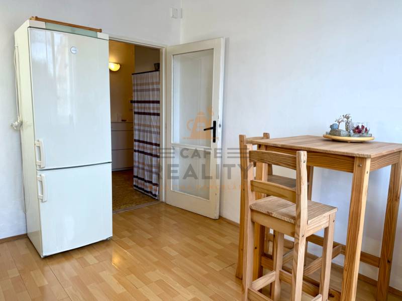 PRENÁJOM 1 izbový byt, 40 M2, Bratislava, Ružinov, Košická ulica, NIVY - Kuchyňa v 1-izbovom byte s dreveným stolom, stoličkami a chladničkou, podlaha s dreveným dekorom.
