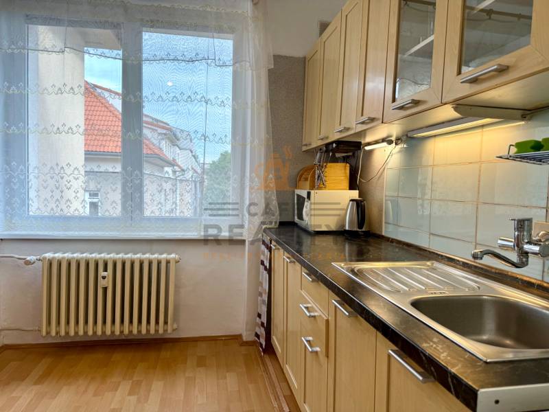 PRENÁJOM 1 izbový byt, 40 M2, Bratislava, Ružinov, Košická ulica, NIVY - Kuchynská linka v 1-izbovom byte s podlahou s dreveným dekorom a výhľadom z okna.