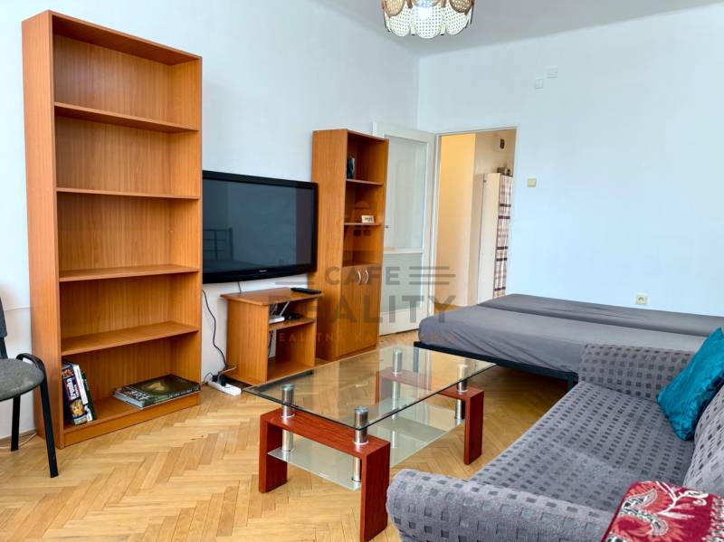 PRENÁJOM 1 izbový byt, 40 M2, Bratislava, Ružinov, Košická ulica, NIVY - Obývacia izba s policou, televízorom, pohovkou a podlahou s dreveným dekorom v 1-izbovom byte.