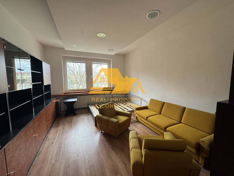 Prenajmem 1 izbový byt v Nových Zámkoch - Obývacia izba v 1-izbovom byte s podlahou s dreveným dekorom, žltou sedačkou a veľkým oknom.