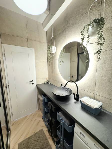 - PRENÁJOM - 3 IZBOVÝ BYT - WILSONOVA UL. - STARÉ MESTO - Kúpeľňa s oblým zrkadlom, závesnými kvetmi a podlahou s dreveným dekorom.