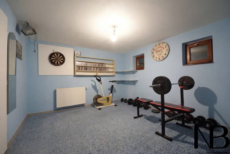 Rodinný dom s veľkým pozemkom a priestorom na podnikanie – T.Jastrabie - Posilňovňa v rodinnom dome s činkami, rotopedom a šípkami na modrej stene.