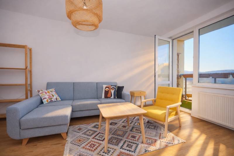 NA PREDAJ-3-izbový byt s loggiou a výhľadom na centrum mesta, KE-JUH - Obývačka so sedačkou, žltým kreslom, koberec a podlaha s dreveným dekorom v 3-izbovom byte.